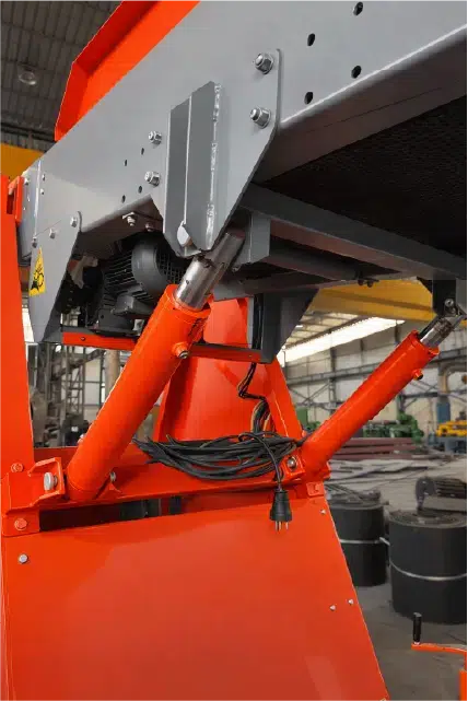 Close-up of an orange conveyor showing hydraulic cylinders and mounting brackets used for height adjustment.