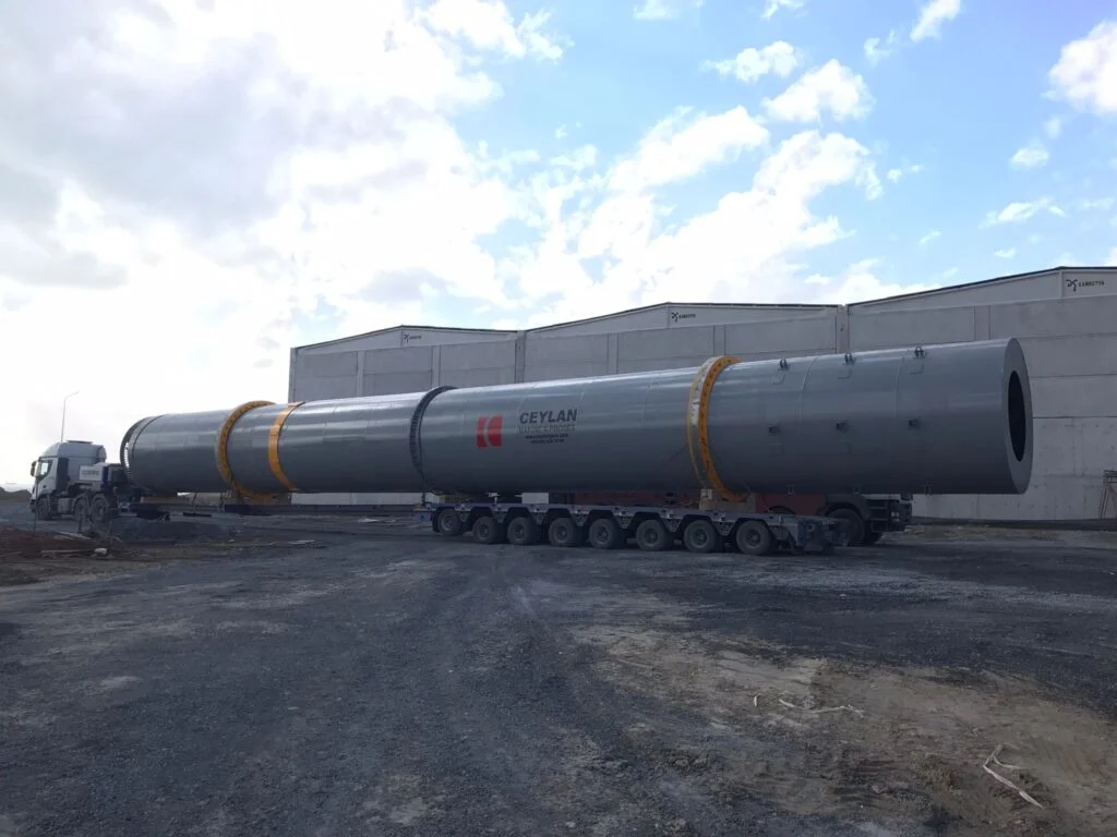 Large industrial rotary drum loaded on a multi-axle trailer outside a factory building